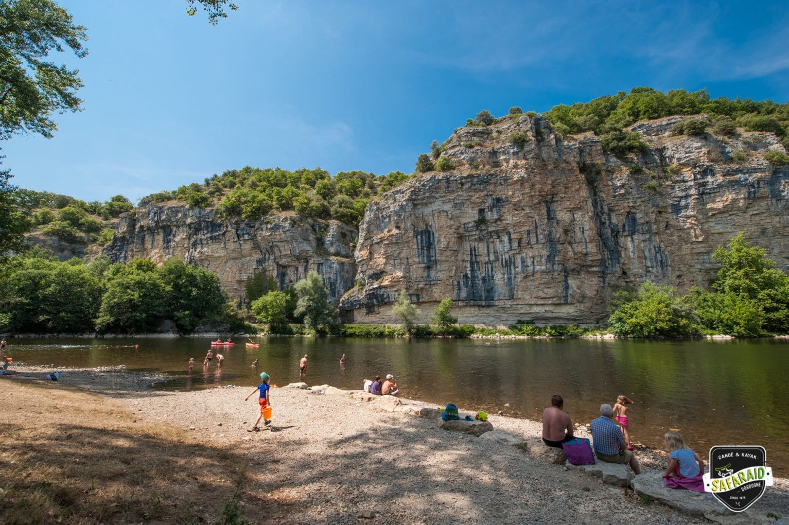 Des familles se baignent dans la rivière sur une plage de galet avec une falaise en face.